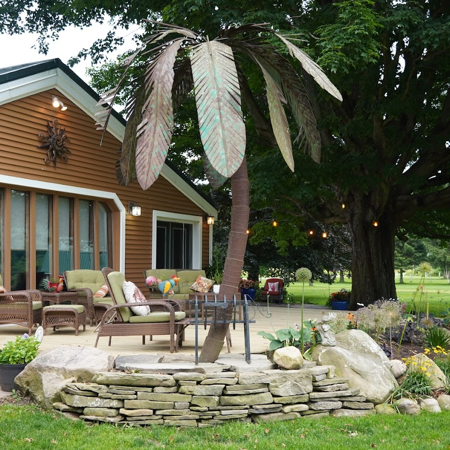 View of the back patio with the metal palm tree and the whicker chairs with green cushions. stone wall frames the area with the big tree in the background. Against the farm house towards the big windows and door