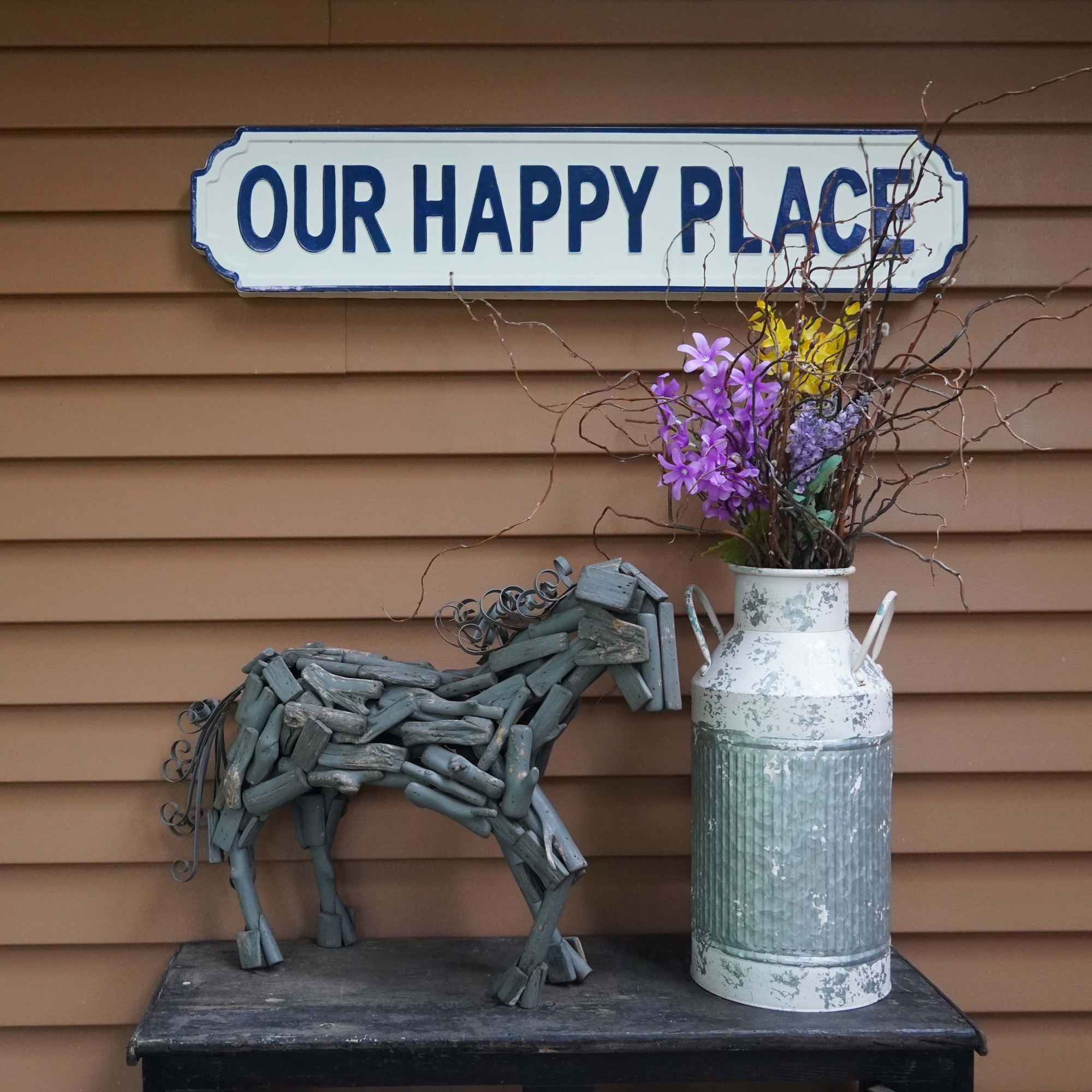 A white and blue sign that reads "Our Happy Place" attached to the siding of the house sitting above a table where a statue of a horse made out of pieces of wood sits next to an old milk can with purple and yellow flowers