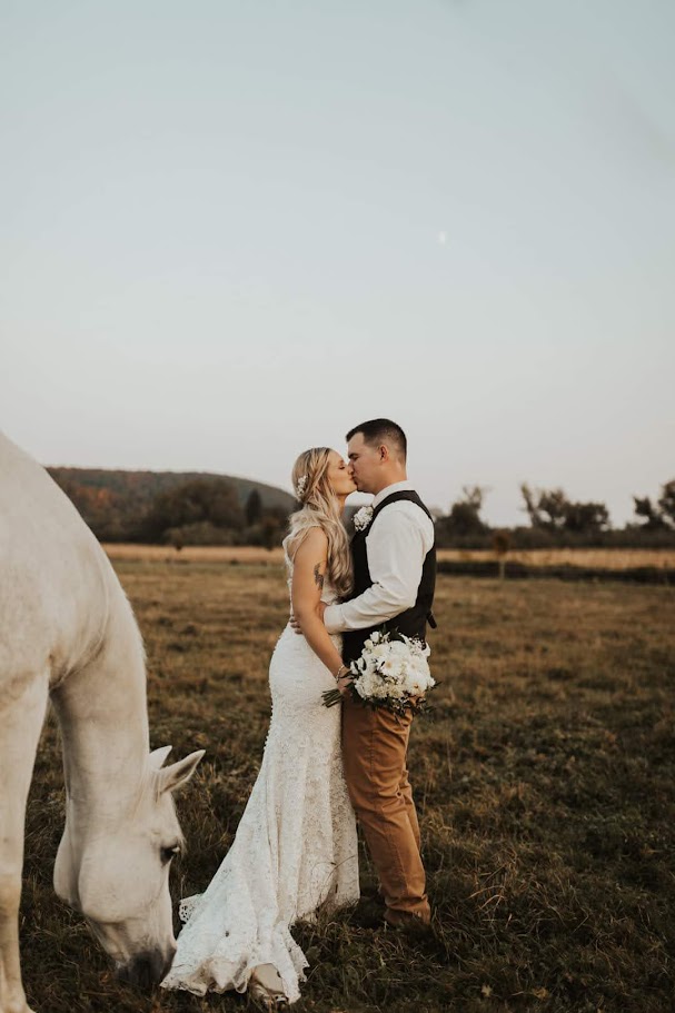 bride and groom sharing a kiss in the field with the white horse bent down next to her gown. bride is holding a white bouqet