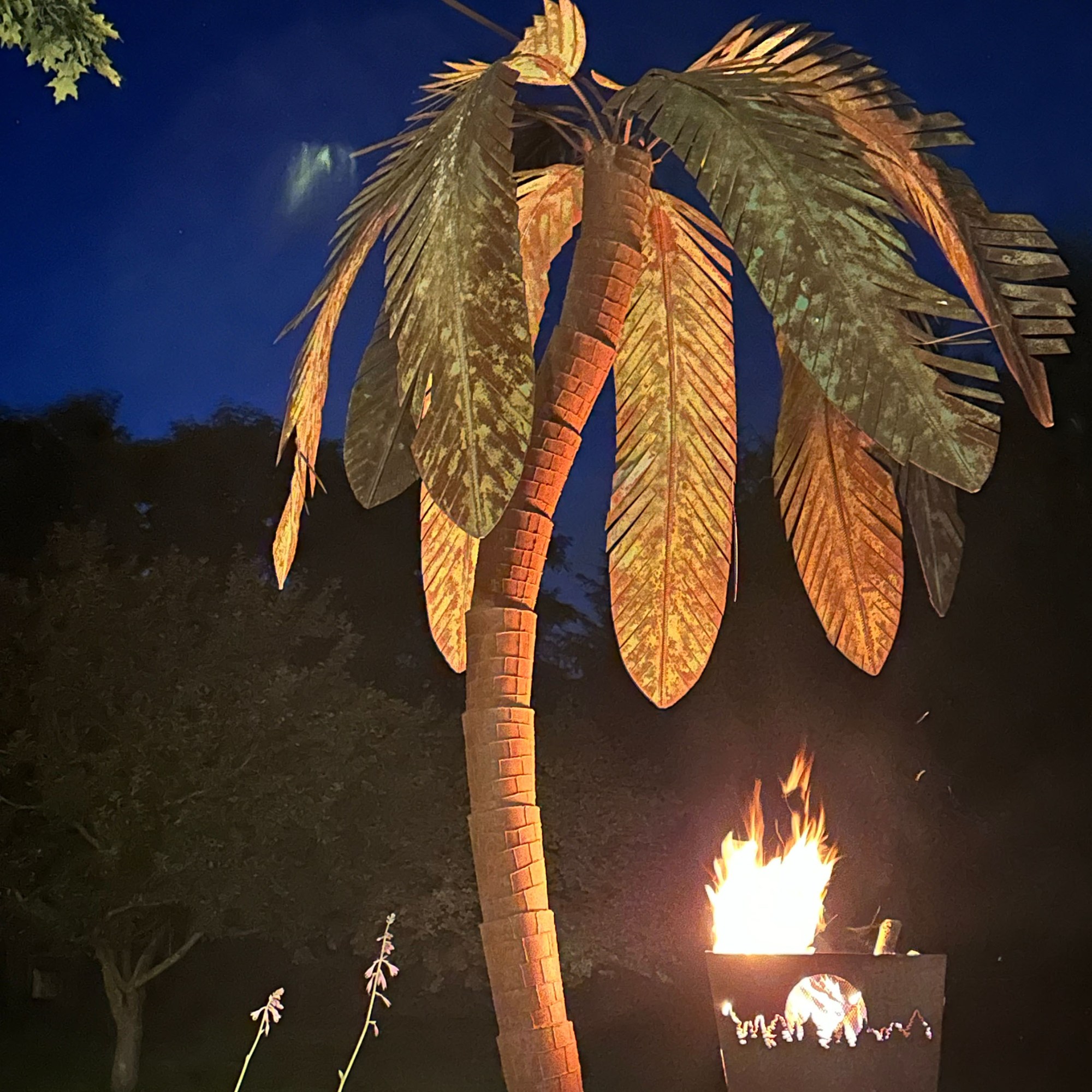 metal palm tree at night with a small fire pit burning to it's right