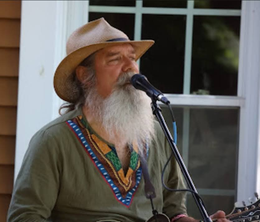 Close up of a man with a tan hat, long white beard, and green shirt playing the guitar and singing
